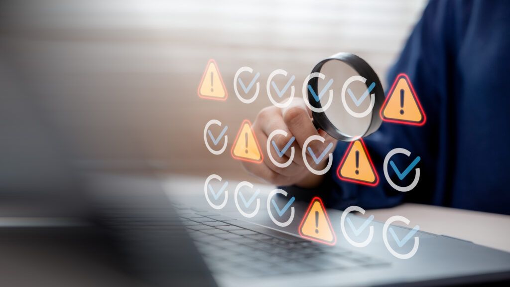 Hand using magnifying glass to inspect warning and check icons on laptop screen, representing network performance monitoring